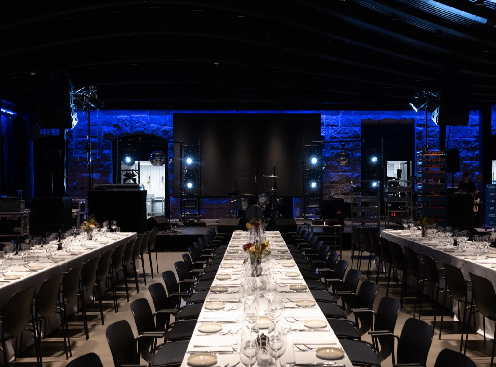 Event space at Villa Copenhagen with long dressed tables, black chairs, a stage with a drum kit and lighting, and blue-lit stone walls